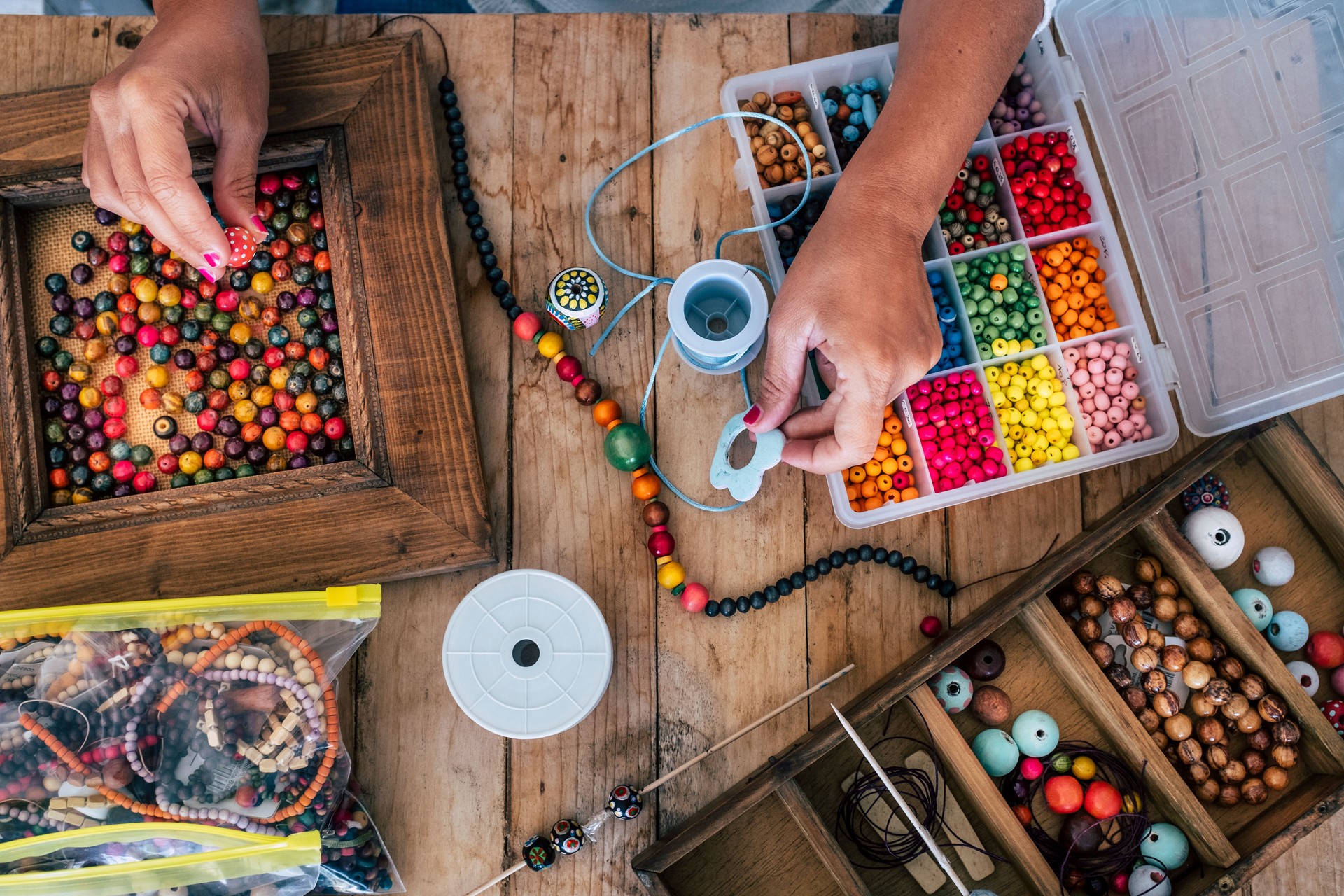 Making of handmade jewellery. Box with beads on old wooden table. Top view with woman hands - tutorial to learn how to make bracelets and jewellery online and alternative home job